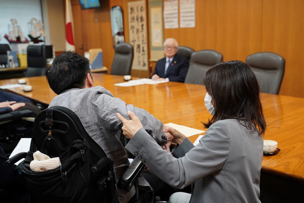 「あ、か、さ、た、な話法」で発言する天畠の後ろ姿。右横で介助者が天畠の腕を引いて読み取っている。机をはさんで正面に武見大臣が着席している。